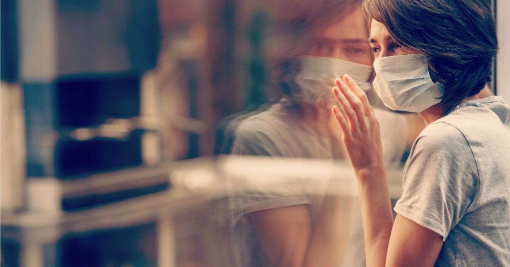A masked woman looks out of her window.