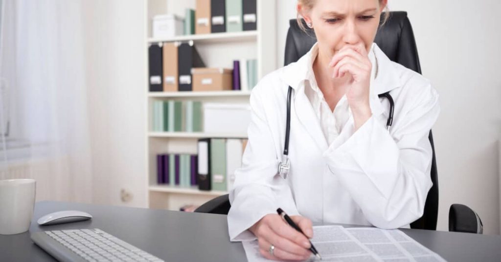 Distressed doctor sitting at at desk, looking at her professional liability insurance policy.