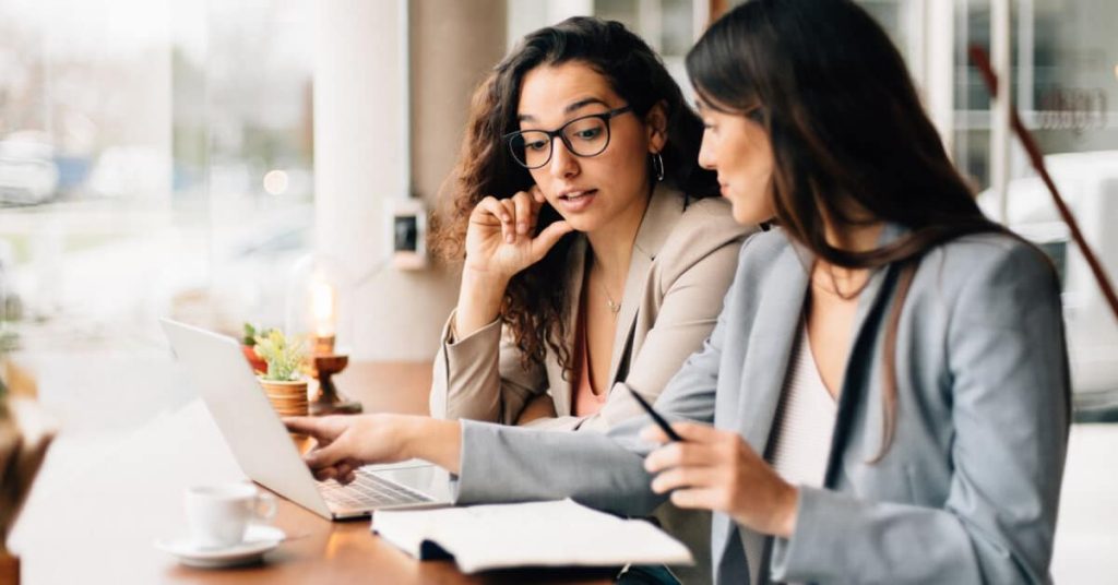 Two professional women sitting at a table, looking at a laptop computer