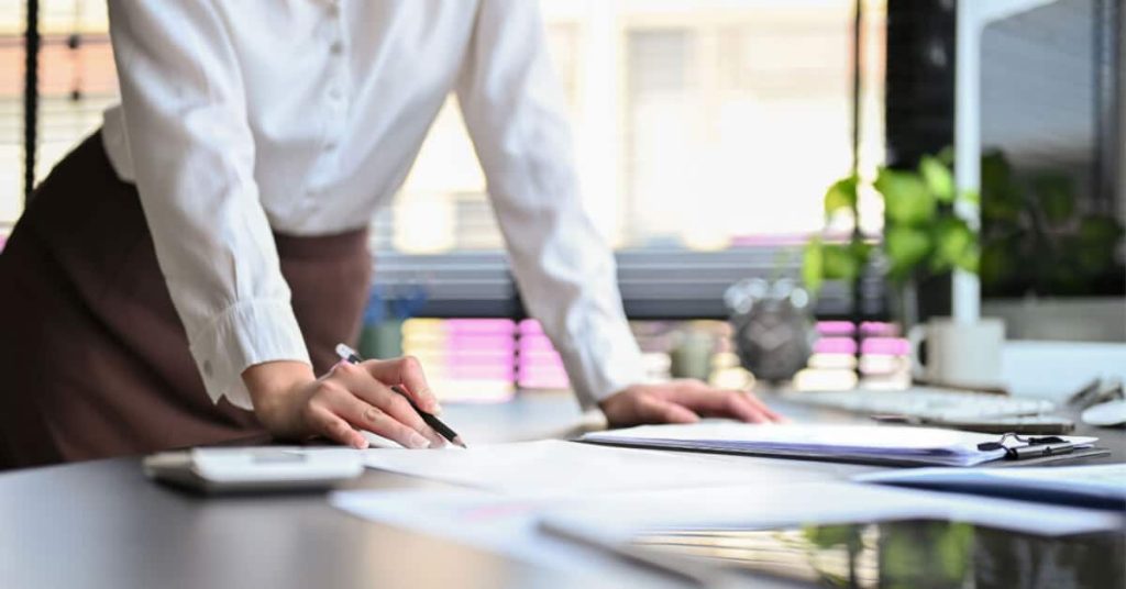 A woman's torso as she leans on a desk with papers.
