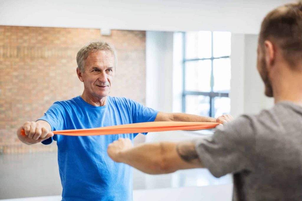 Man doing physical therapy exercises  with a band under the supervision of a trainer.