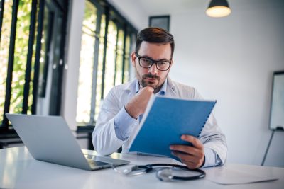 Physician at his desk reviews a bound study protocol, considering whether his small practice can run a clinical trial.