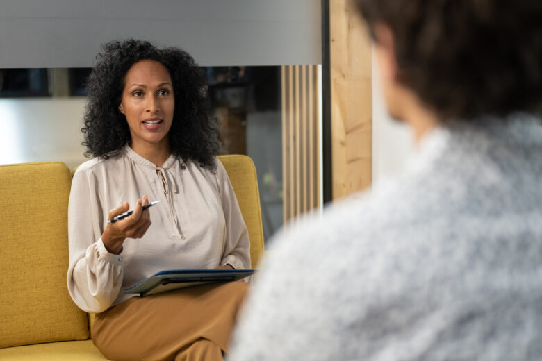 A therapist holding a notepad and pen speaks with a client during a session about her missed appointment fee policy.