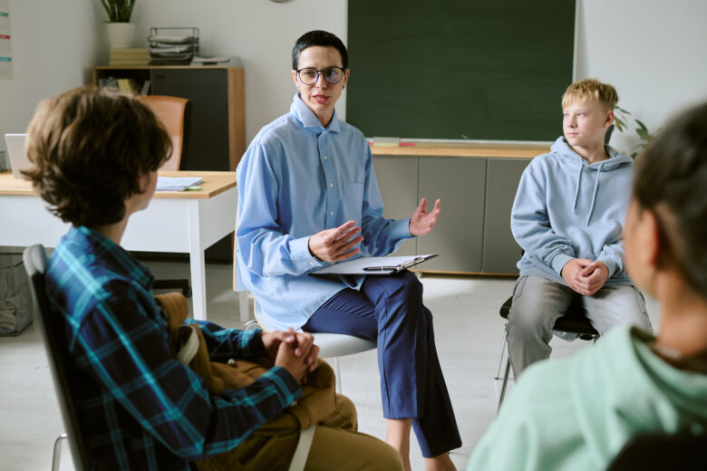 Adult counselor holding a clipboard leads a group therapy session with teenagers seated in a circle at a youth IOP or PHP behavioral health program.